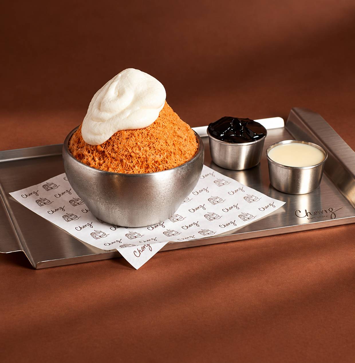 Steel bowl of orange shaved ice with a swirl of cream, beside small cups of grass jelly and condensed milk.