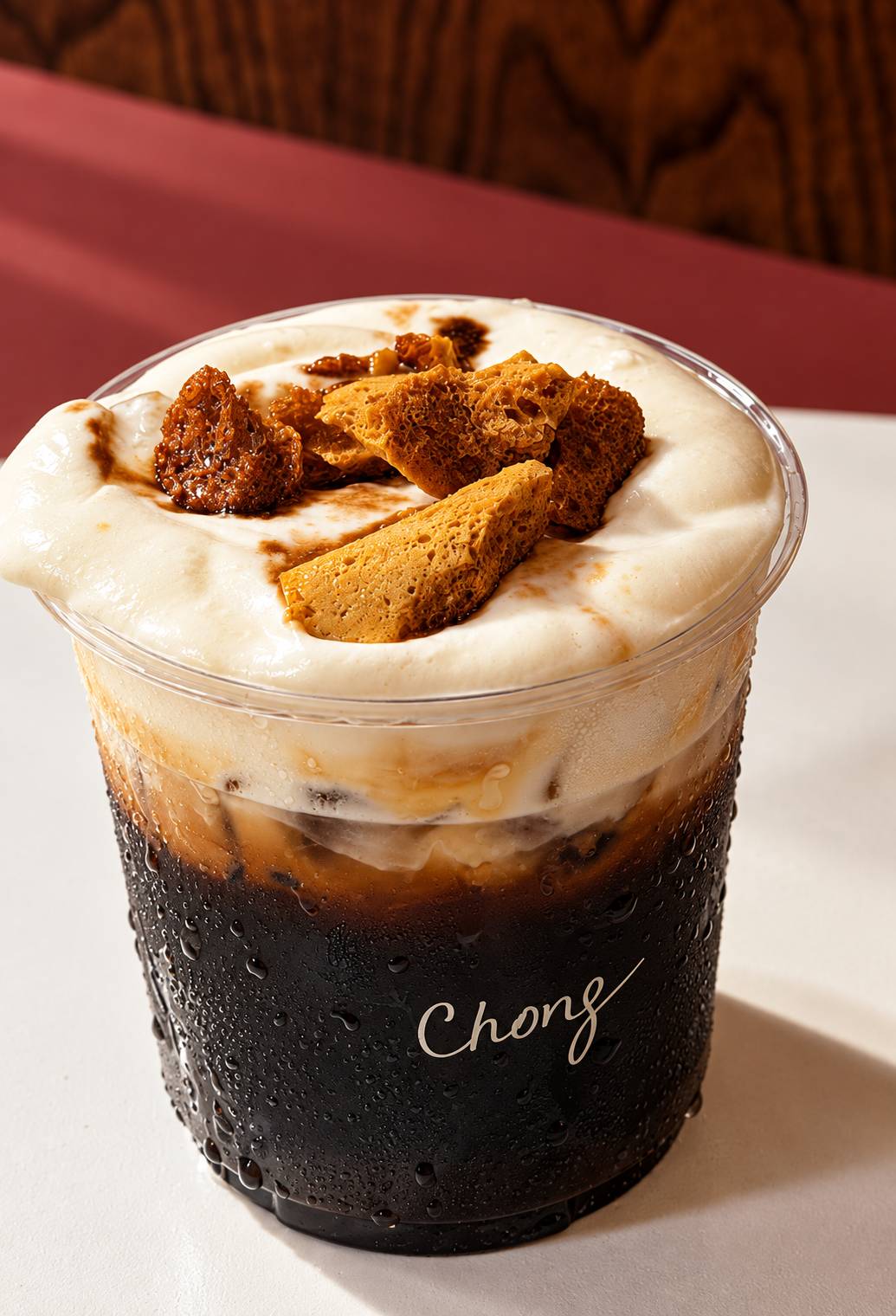 Dark iced drink topped with cream foam and pieces of honeycomb, in a cup marked Chong.