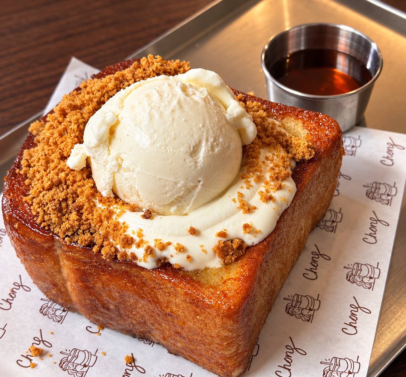 Thick golden toast topped with a scoop of ice cream, cream and crumble, beside a small cup of syrup.