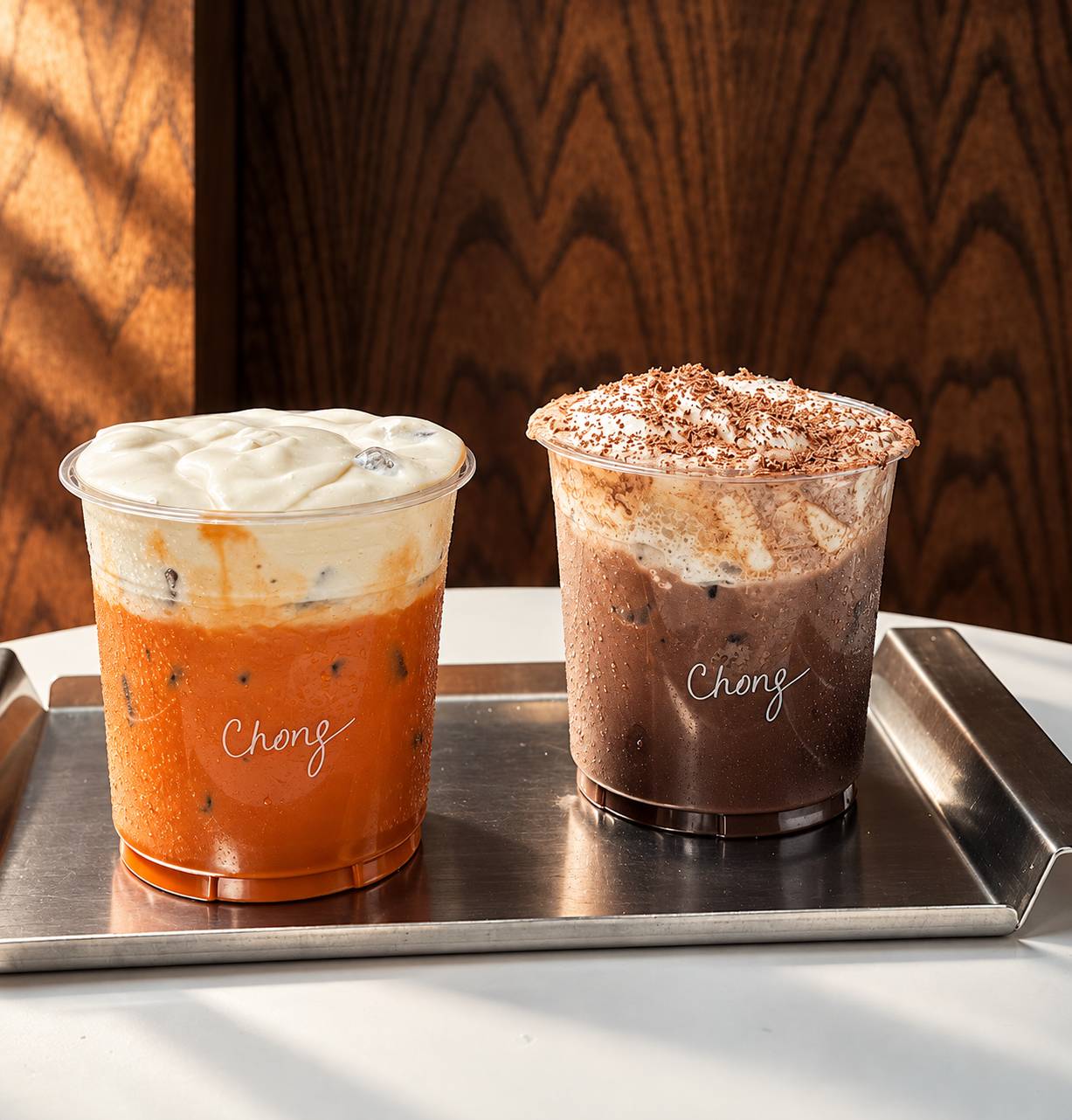 Two iced drinks on a steel tray: an orange tea with cream foam and a cocoa drink with shaved chocolate.